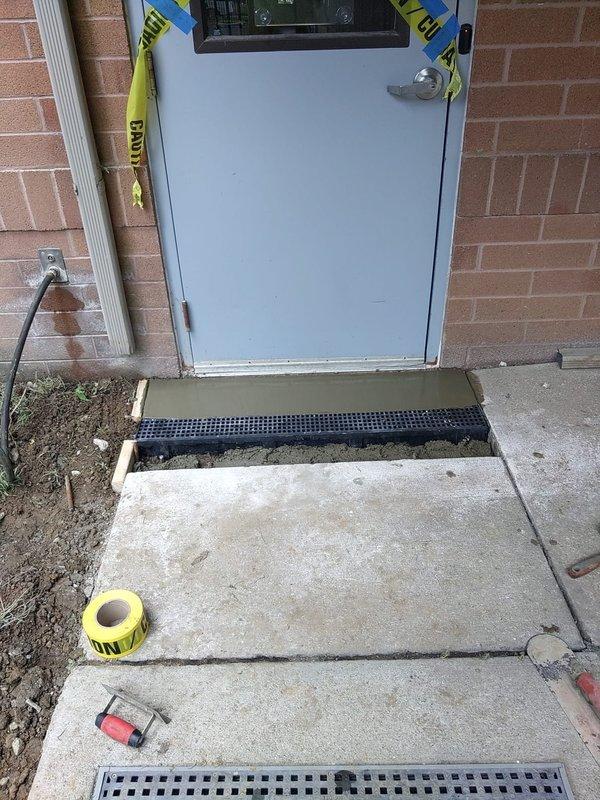 Installed trench drain system at building exterior doorway entrance per specifications. The drainage channel with black perforated grate was set into freshly poured concrete at the threshold to manage water runoff and prevent pooling at the entryway. Work area was secured with caution tape during concrete curing process.