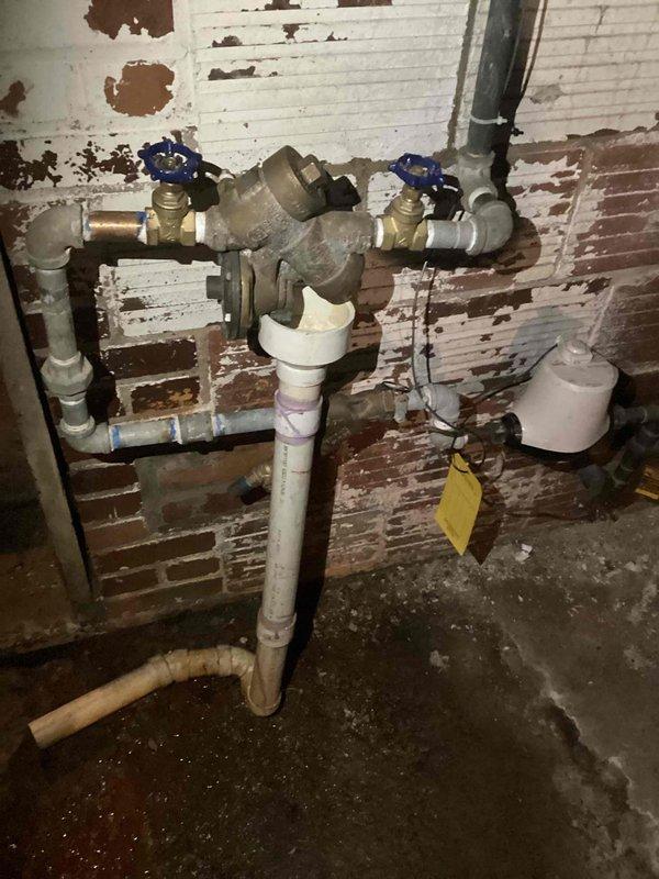 Responded to service call for backflow preventer evaluation. Upon arrival, located backflow assembly mounted on brick wall in basement utility area. Inspection revealed backflow prevention device had been previously tampered with by unauthorized repair attempt, resulting in compromised functionality and integrity of the assembly. Device showed signs of improper installation work and could not be restored to proper working condition. 

Documented existing plumbing configuration consisting of brass backflow preventer connected to copper and PVC piping with blue-handled shut-off valves positioned on either side of the assembly. Photographed complete installation including vertical and horizontal pipe runs, valve placements, and overall condition of the assembly for quote preparation. 

Determined backflow preventer requires complete replacement due to failed repair attempt and inability to restore device to code-compliant operational status. Advised customer that replacement of entire bac