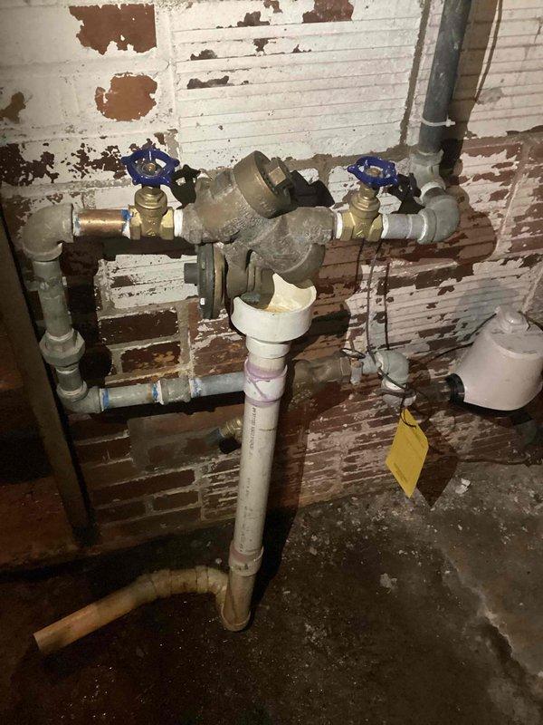 Responded to service call for backflow preventer evaluation. Upon arrival, located backflow assembly mounted on brick wall in basement utility area. Inspection revealed backflow prevention device had been previously tampered with by unauthorized repair attempt, resulting in compromised functionality and integrity of the assembly. Device showed signs of improper installation work and could not be restored to proper working condition. 

Documented existing plumbing configuration consisting of brass backflow preventer connected to copper and PVC piping with blue-handled shut-off valves positioned on either side of the assembly. Photographed complete installation including vertical and horizontal pipe runs, valve placements, and overall condition of the assembly for quote preparation. 

Determined backflow preventer requires complete replacement due to failed repair attempt and inability to restore device to code-compliant operational status. Advised customer that replacement of entire bac