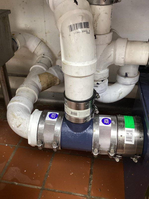 Responded to commercial property for dishwasher drain obstruction. Upon inspection, located clogged drain line in under-sink plumbing assembly consisting of white PVC piping with flexible coupling connection secured by metal hose clamps. Successfully cleared blockage from dishwasher drain system, restoring proper drainage function. Verified system operation and confirmed water flow through drain assembly without backup or restriction.