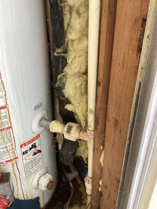 Conducted inspection of Richmond 6-year warranty water heater in utility space showing signs of age with corrosion on pipes and connections. Noted installation in confined area surrounded by fiberglass insulation with inadequate clearance and visible cobwebs. Documented cold water inlet connection and PVC piping configuration for replacement estimate.
