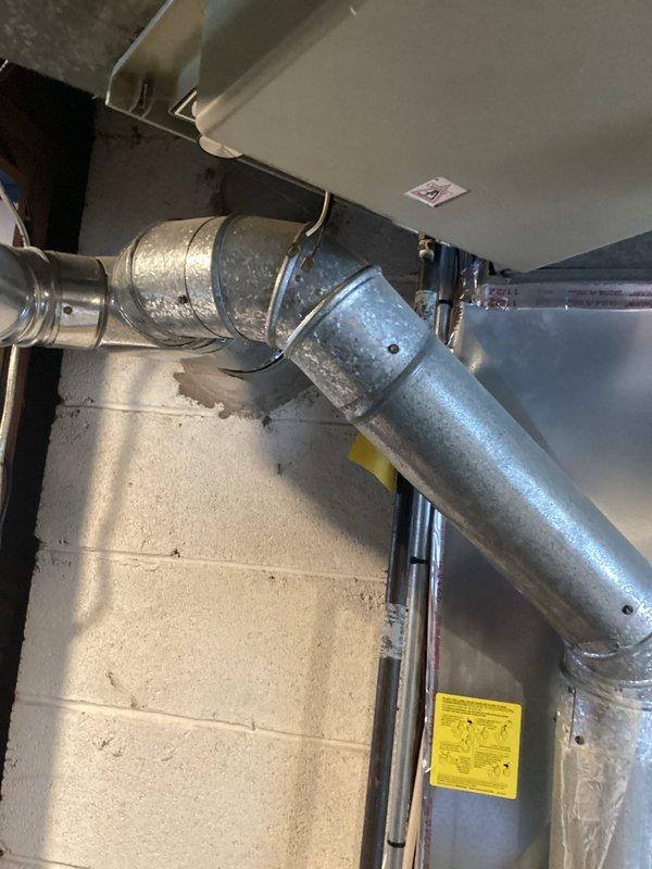 Performed assessment of aging HVAC system (9-11 years old) for potential replacement. Inspected existing ductwork configuration, including galvanized steel ducts with elbow joints mounted along concrete block wall. Evaluated condensate drainage system with white drain box and associated piping. Documentation included photographs of critical system components to support replacement recommendation due to equipment age.