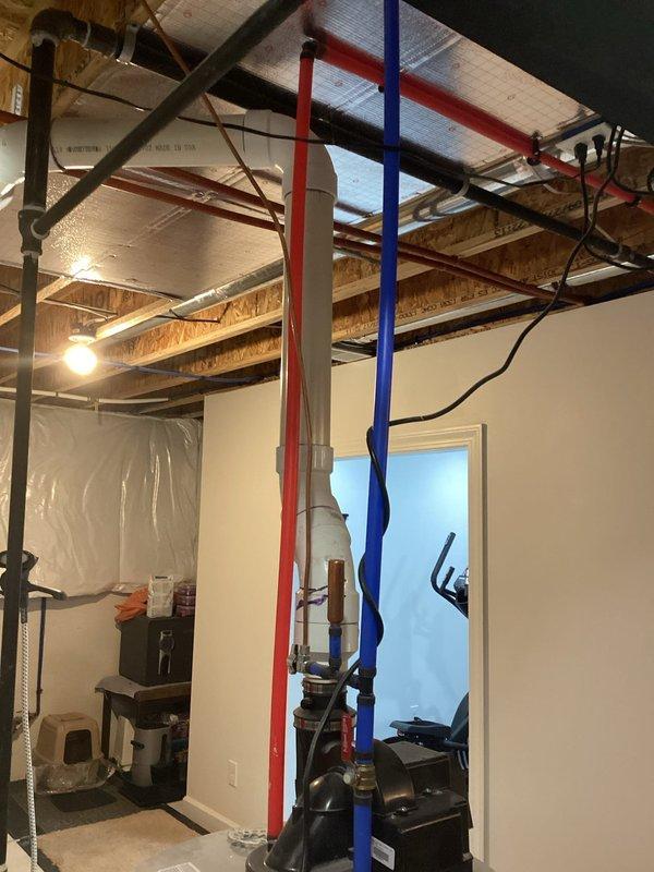 Conducted water heater estimation in unfinished basement utility area featuring exposed ceiling joists with extensive plumbing infrastructure including red/blue supply lines and PVC drainage pipes. Assessment included evaluation of current installation conditions, space requirements, and necessary connections for proper replacement, with photo documentation taken of the mechanical room setup for accurate quote preparation.