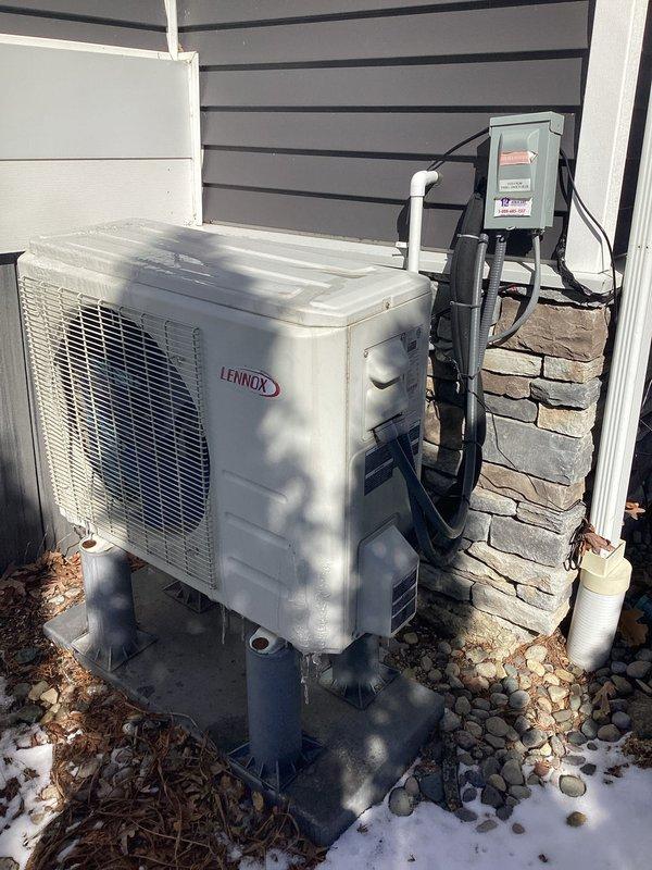 Completed estimate for mini-split installation featuring an interior wall-mounted Lennox ductless air handler in client's cabin-style room and corresponding exterior condenser unit mounted on metal support legs with proper electrical connections. System was properly installed with line sets routed through exterior wall, with condenser positioned on decorative river rock landscaping area.