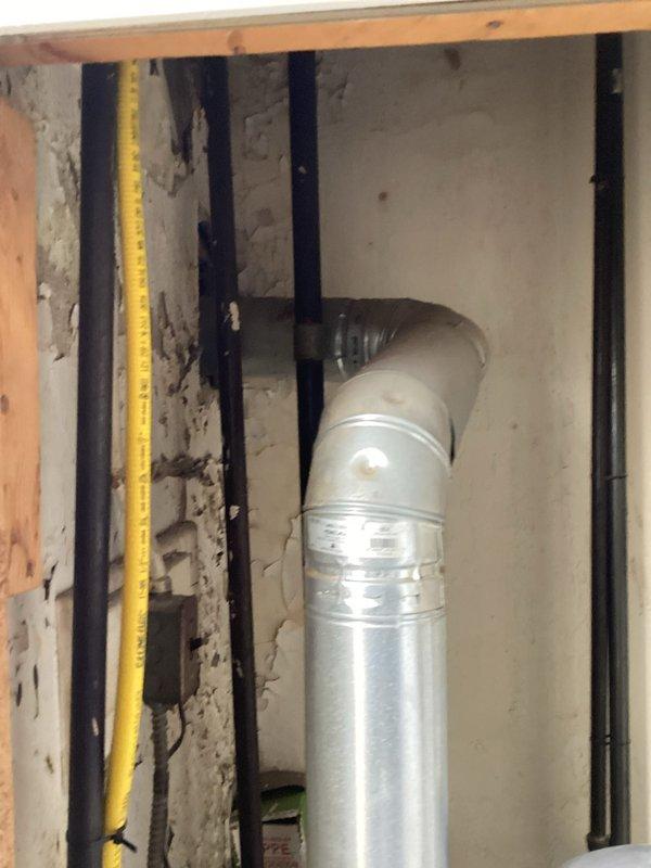 Conducted boiler assessment in commercial building's utility room, documenting significant wall deterioration around existing hot water system. Inspection revealed aging boiler components including deteriorated flue pipe, expansion tank connections, and gas line installation. Photographed exposed ductwork and framing requiring attention during potential replacement. Recommended comprehensive system upgrade due to extensive structural damage and aged equipment condition.