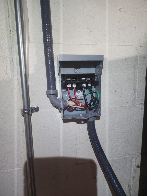 Installed electrical disconnect box on hybrid water heater per code requirements. Mounted gray metal disconnect enclosure on concrete block wall adjacent to unit, ran metallic and liquid-tight flexible conduit from disconnect to water heater terminals, and completed all wiring connections with proper color-coded conductors to terminal blocks.