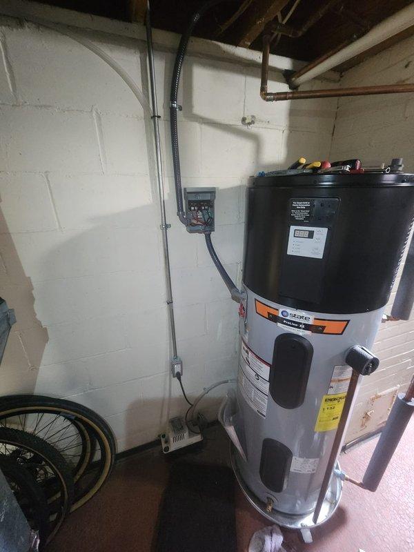 Installed electrical disconnect box on hybrid water heater per code requirements. Mounted gray metal disconnect enclosure on concrete block wall adjacent to unit, ran metallic and liquid-tight flexible conduit from disconnect to water heater terminals, and completed all wiring connections with proper color-coded conductors to terminal blocks.