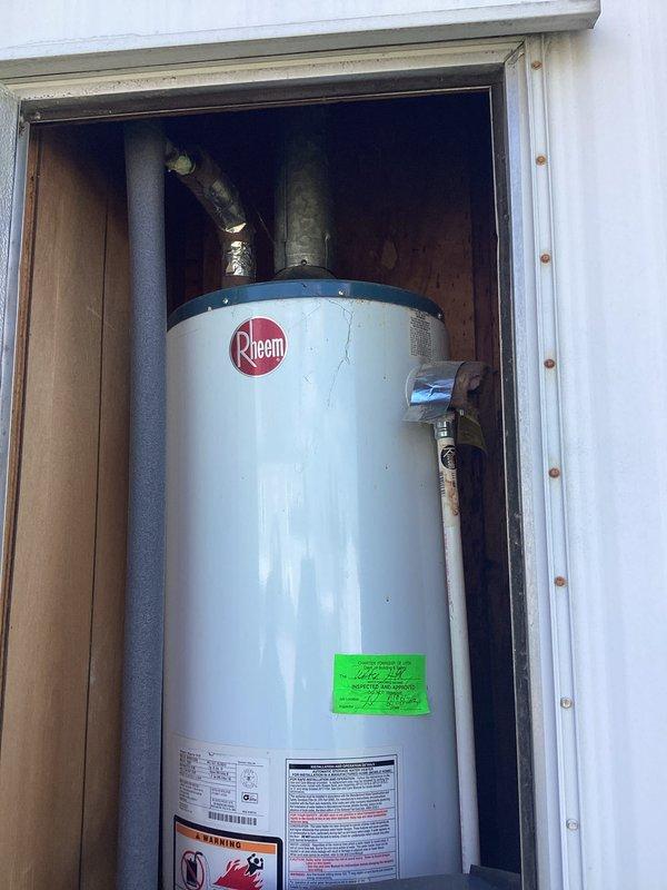 Provided comprehensive estimate for complete water heater replacement of existing Rheem residential unit currently installed in exterior access compartment with corrugated metal siding. Evaluated current installation featuring white cylindrical water heater with visible age-related deterioration on plumbing connections, including weathered bare metal piping and black foam-insulated lines. Assessment included inspection of yellow flexible gas line connector, temperature control valve, and all associated plumbing fixtures within wooden-framed utility compartment. Documented existing conditions through photographic record for replacement specifications and scope of work development.