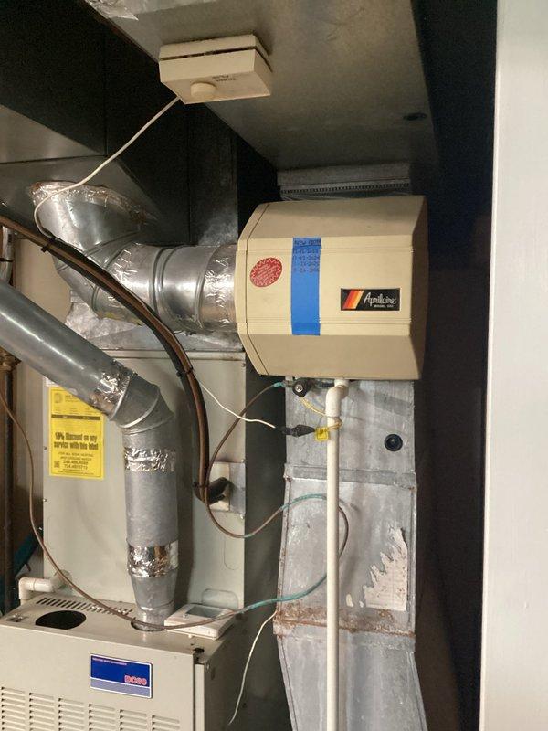 Conducted comprehensive evaluation of 27-year-old residential furnace system experiencing control board failure. Assessed existing equipment configuration including furnace unit, ductwork connections, humidifier integration, and associated refrigerant lines. Documented current system condition through detailed photography showing white cabinet-style furnace with ventilated front panel, gray metal ductwork with foil tape joints, wall-mounted humidifier unit with water line connections, and copper refrigerant piping. Provided replacement estimate options for complete HVAC system upgrade due to age of equipment and failed control board component.