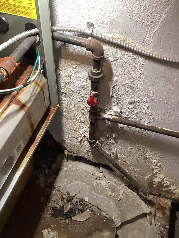 # HVAC Service - Water Leak

Responded to service call for furnace leaking from bottom in confined installation space. Unit is an 11-year-old system manufactured in 2015, currently operational and providing heat. Upon inspection, discovered significant rust staining and corrosion on interior metal panels and surfaces, indicating ongoing moisture intrusion. Heavy vertical rust streaking observed on internal components, suggesting water dripping from upper sections of unit. 

Examined condensate drainage system and identified issues with condensate removal. The copper piping and connections showed advanced oxidation and reddish-brown corrosion, particularly on lower sections. Vertical pipe assembly includes combination of copper and black iron pipe with visible deterioration. Evidence of prolonged water exposure present throughout mechanical compartment.

Utility area shows aging infrastructure with corroded metal components, cracked concrete flooring, and deteriorated wall surfaces at b