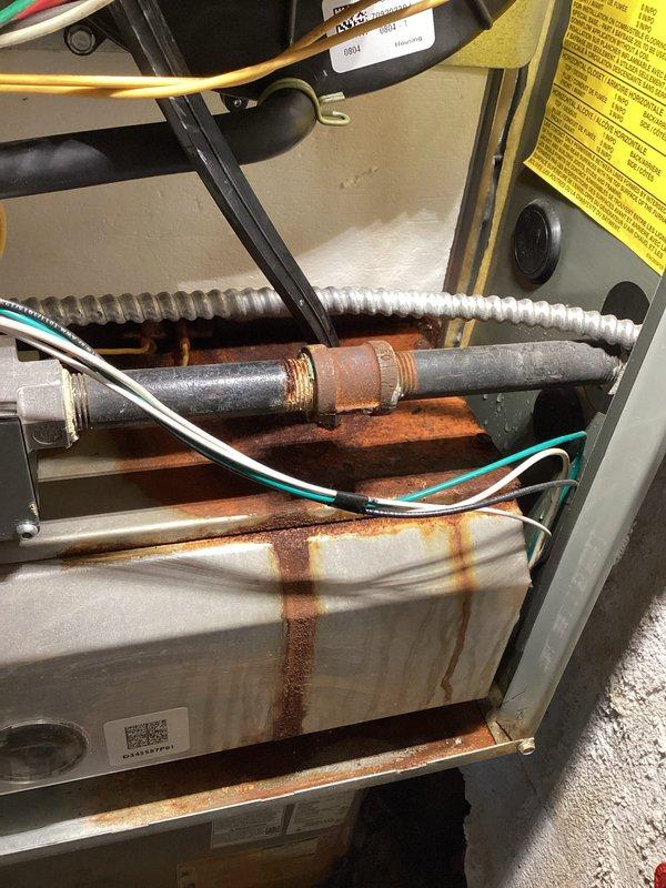 # HVAC Service - Water Leak

Responded to service call for furnace leaking from bottom in confined installation space. Unit is an 11-year-old system manufactured in 2015, currently operational and providing heat. Upon inspection, discovered significant rust staining and corrosion on interior metal panels and surfaces, indicating ongoing moisture intrusion. Heavy vertical rust streaking observed on internal components, suggesting water dripping from upper sections of unit. 

Examined condensate drainage system and identified issues with condensate removal. The copper piping and connections showed advanced oxidation and reddish-brown corrosion, particularly on lower sections. Vertical pipe assembly includes combination of copper and black iron pipe with visible deterioration. Evidence of prolonged water exposure present throughout mechanical compartment.

Utility area shows aging infrastructure with corroded metal components, cracked concrete flooring, and deteriorated wall surfaces at b