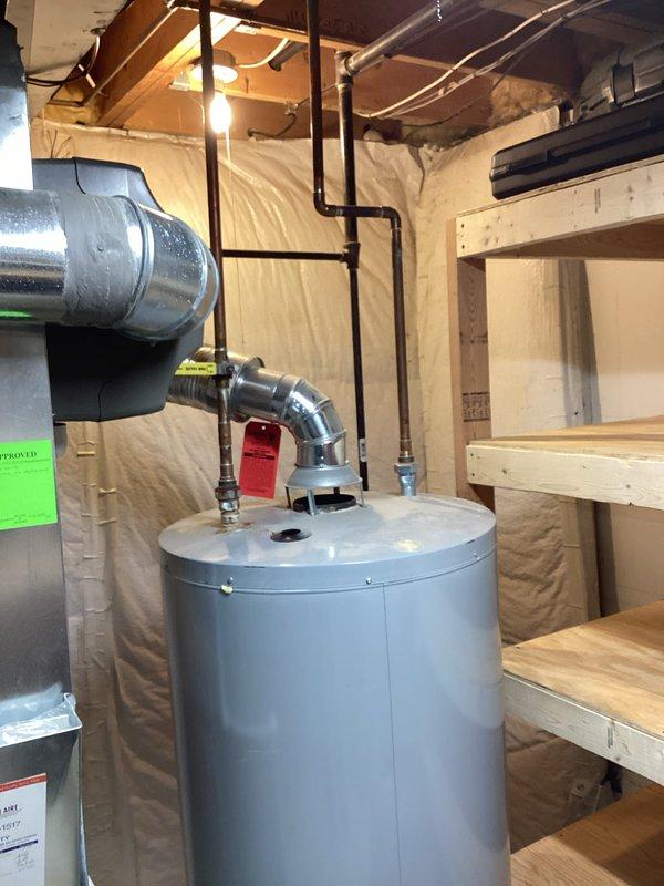 Responded to customer request for water heater replacement estimate due to reported leaking unit. Upon arrival, assessed existing cylindrical water heater located in unfinished basement utility area with exposed joists and insulation. Documented current installation including all pipe connections, copper supply lines, red-handled shutoff valve, and top-mounted fittings. Evaluated surrounding plumbing infrastructure including PVC drainage system and gas line configuration. Photographed unit positioning relative to HVAC ductwork and available clearances for replacement access. Provided comprehensive estimate for complete water heater replacement including removal of existing unit and installation of new equipment with proper venting and code-compliant connections.