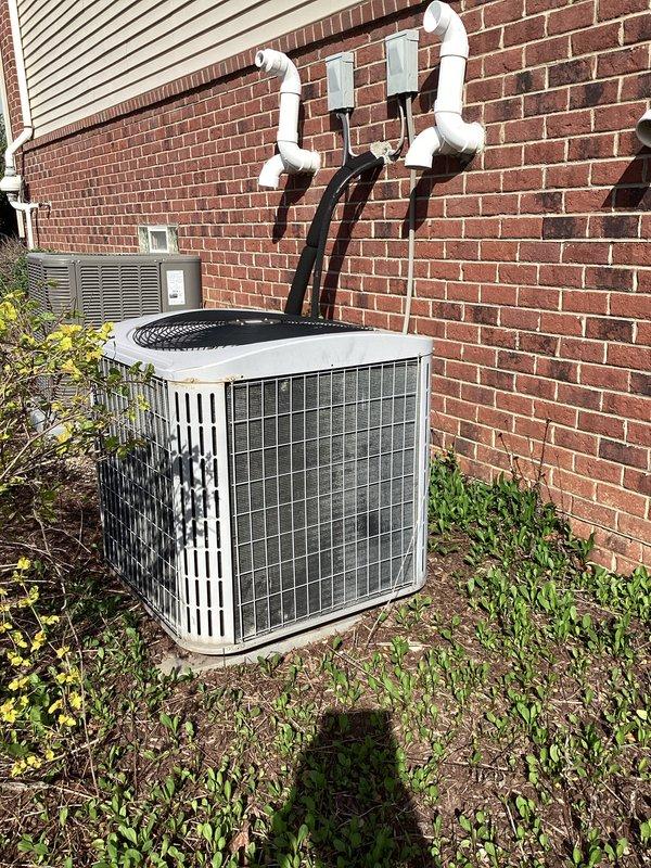 Conducted comprehensive on-site evaluation for complete HVAC system replacement at residential property. Assessed existing Carrier indoor air handler and furnace unit located in basement utility area, along with outdoor condensing unit positioned against exterior brick wall. Documented current system configuration including multi-zone split system setup with multiple refrigerant line sets penetrating building envelope. Photographed indoor equipment installation showing metal cabinet units with connecting ductwork in unfinished basement space with exposed framing. Evaluated outdoor condenser unit condition, noting white/beige cabinet with protective grilles, existing electrical disconnect box, and refrigerant line routing through brick wall penetrations. Captured images of installation environment including PVC drainage piping, flexible ductwork connections, and equipment placement relative to structural elements. Provided detailed photographic documentation of both indoor and outdoor c