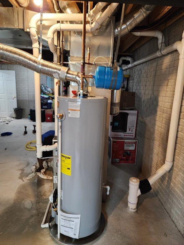 Completed warranty replacement installation of residential tanked water heater in basement mechanical room. New unit installed alongside existing Bryant furnace system with proper connections to water supply lines, gas line, and venting through white PVC piping. Blue expansion tank mounted and integrated into system per code requirements, all copper piping connections secured and tested for leaks, installation verified operational.