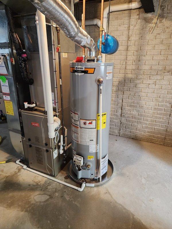 Completed warranty replacement installation of residential tanked water heater in basement mechanical room. New unit installed alongside existing Bryant furnace system with proper connections to water supply lines, gas line, and venting through white PVC piping. Blue expansion tank mounted and integrated into system per code requirements, all copper piping connections secured and tested for leaks, installation verified operational.