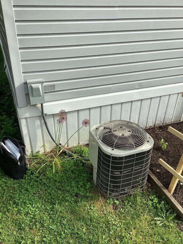 Responded to service call for non-functional air conditioning system at mobile home residence. Evaluated existing outdoor condenser unit of undetermined age with cylindrical design and beige casing, noting furnace had been previously replaced while AC unit remained original. Unit located on ground level adjacent to house siding with electrical disconnect box and conduit visible. Documented equipment condition and installation site through photographs showing condenser positioning, electrical connections, and surrounding landscaping. Provided customer with estimate for complete AC system replacement to restore cooling functionality.