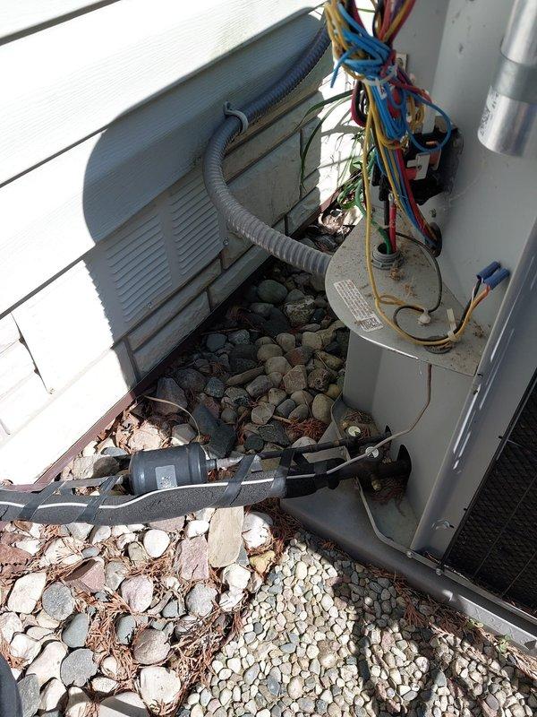 # HVAC Service Call - No Cooling Condition

Responded to service call for residential heat pump system reporting no cooling operation. Upon arrival, performed visual inspection of outdoor condensing unit installed on exterior wall with river rock landscaping base. Unit exhibited signs of age with visible corrosion on mounting brackets and oxidation on metal surfaces consistent with 10+ year service life.

Conducted diagnostic evaluation of electrical connections and control components. Observed multiple conductor wiring entering unit through side panel with proper conduit installation. Serial number documented from equipment nameplate for service records. Noted accumulation of organic debris including leaves and pine needles around unit base which can restrict airflow and reduce system efficiency.

Assessed overall system condition including condensing coil fins, electrical terminations, and refrigerant line connections. Unit displayed typical wear patterns associated with extended out