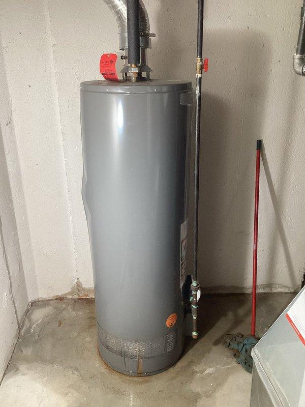 Responded to service call for leaking water heater in unfinished basement with walk-out access. Inspected 10-year-old residential water heater unit showing signs of active leak at base, with visible moisture staining on concrete floor. Documented existing installation including top-mounted plumbing connections, pressure relief valve, copper supply lines, flexible vent ductwork, and safety strapping. Photographed unit specifications, manufacturer labels, and overall condition to prepare replacement estimate as requested.