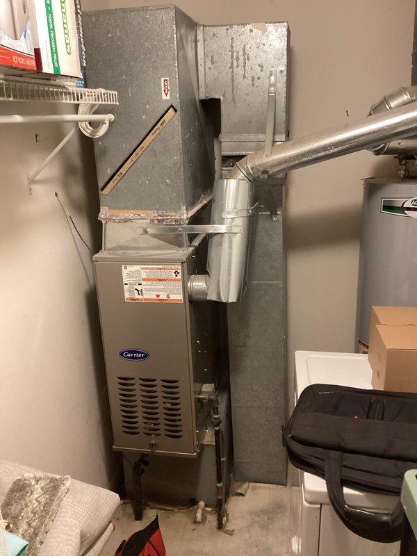 Conducted on-site assessment of existing air conditioning system to provide estimate for unit relocation from side yard to rear of property to meet updated municipal code requirements. Evaluated older model outdoor condenser unit showing weathering and vegetation encroachment, with age undetermined as system was present at time of home purchase. Inspected indoor Carrier furnace and ductwork configuration in utility area to determine feasibility of refrigerant line rerouting and electrical modifications required for relocation project. Documented current equipment condition and installation layout with photographs for estimate preparation, noting system currently operational with no reported performance issues.
