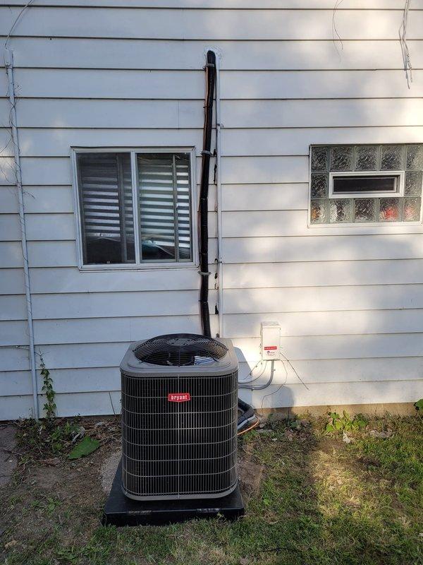 Completed installation of Bryant air conditioning system with downflow configuration at residential property. Installed new condensing unit on exterior platform pad with proper leveling and positioning. Replaced existing lineset with new refrigerant lines, routing copper piping and insulation along exterior wall with secure mounting brackets. Connected gray insulated refrigerant lines from condenser to interior air handler. Installed gray electrical disconnect box on exterior wall per code requirements. Mounted and programmed Ecobee6 smart thermostat to control new system. Ran condensate drain line using white PVC piping from air handler to exterior discharge point. Verified all electrical connections, tested system operation through complete cooling cycle, and confirmed proper refrigerant charge and airflow. System commissioned and left in fully operational condition.