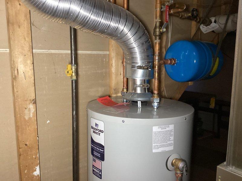 Completed installation of a 40-gallon Bradford White residential water heater in utility area. Installation included proper venting with metallic flex duct, connection of copper supply lines with brass fittings and shut-off valves, installation of blue expansion tank, yellow-handled gas valve, pressure relief valve, and PVC drain pan piping system connected to floor drain.