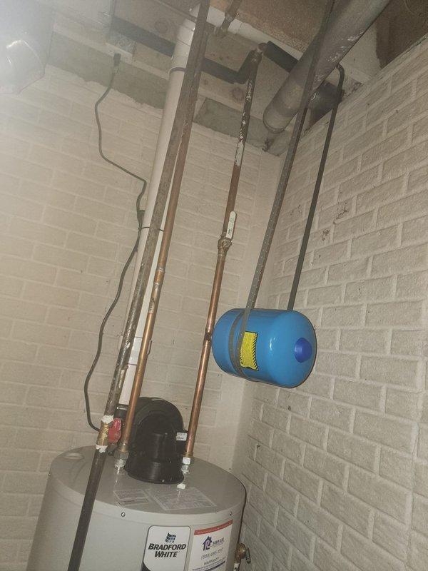 Completed installation of a 50-gallon Bradford White tanked water heater in utility room with white painted brick walls. Connected copper supply lines, installed blue expansion tank suspended from ceiling with metal straps, and properly secured all plumbing components to wall using brackets and supports per manufacturer specifications and local code requirements.