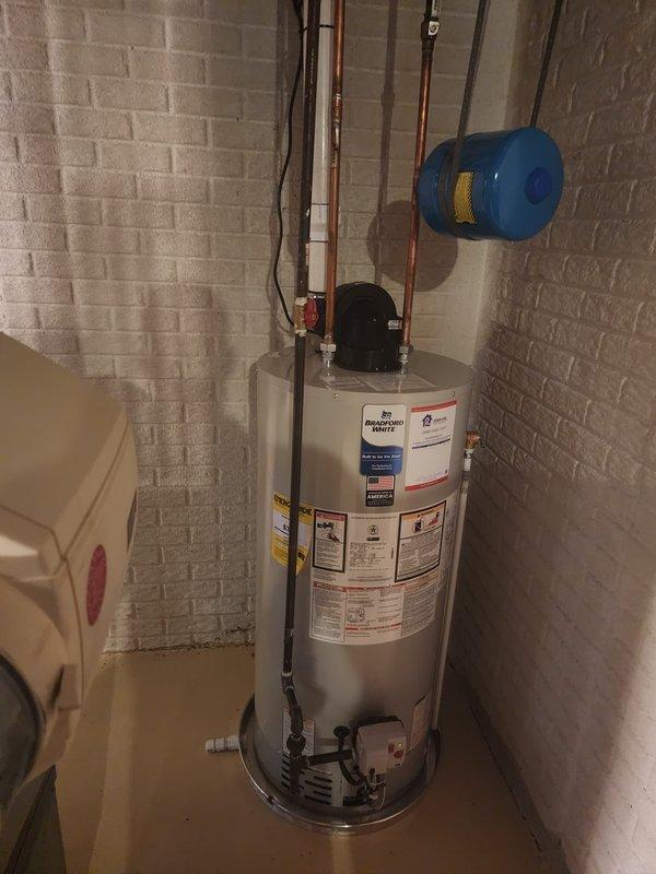Completed installation of a 50-gallon Bradford White tanked water heater in utility room with white painted brick walls. Connected copper supply lines, installed blue expansion tank suspended from ceiling with metal straps, and properly secured all plumbing components to wall using brackets and supports per manufacturer specifications and local code requirements.