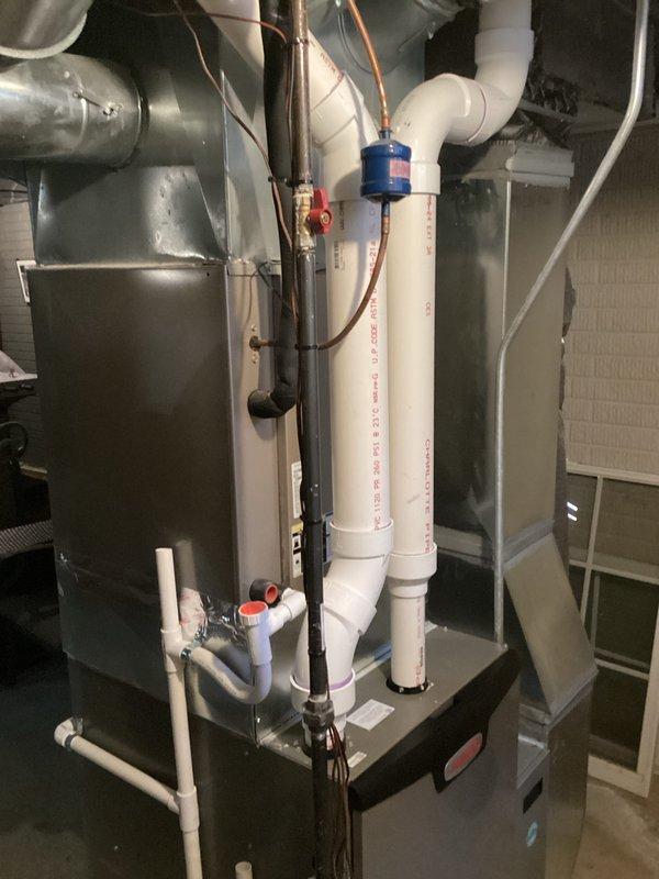 Completed HVAC system replacement including installation of new air handler unit with associated refrigerant lines, condensate drainage system, and ductwork connections. The installation features properly routed copper refrigerant lines, white PVC condensate piping with appropriate fittings and valves, filter drier component, and integrated metallic ductwork for air distribution. All components were installed in the mechanical room with proper clearances and connections verified for system operation.