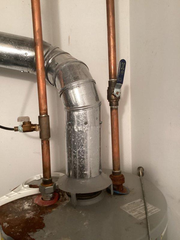 Completed on-site assessment of aging water heater system exhibiting active leakage. Documented existing installation including corroded cylindrical expansion tank or pressure vessel mounted atop water heater, copper supply lines with shutoff valves, and galvanized venting components showing mineral deposits and moisture damage. Photographed current plumbing configuration and identified failed components requiring replacement to provide accurate estimate for new water heater installation.