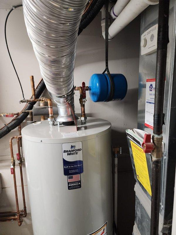 Installed Bradford White tanked water heater in residential utility closet, including all necessary copper supply line connections with shut-off valves, proper venting configuration, and blue expansion tank mounted to ceiling structure. All plumbing connections were completed to code specifications, with the unit positioned for optimal access alongside existing HVAC equipment, ensuring proper clearances and integration with existing PVC drainage and ventilation systems.