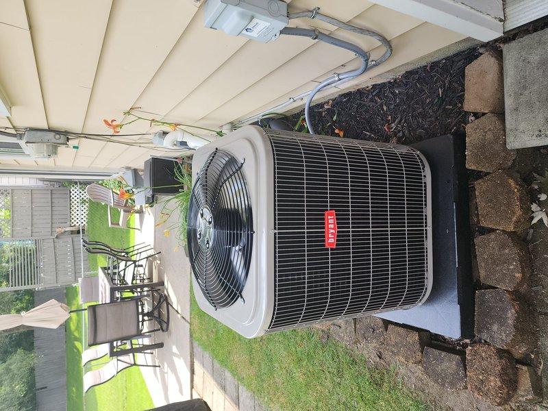 Completed installation of Bryant brand air conditioning condenser unit at residential property. Unit was positioned on concrete pad adjacent to building exterior beneath covered porch area, with all electrical connections, refrigerant lines, and condensate drainage properly installed and tested for optimal system performance.