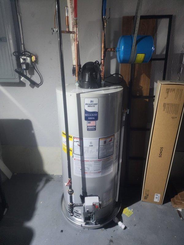 Completed installation of a 50-gallon Bradford White automatic storage natural gas water heater in residential utility room. Connected copper supply lines, installed pressure relief valve, and mounted blue expansion tank to wall. All plumbing connections secured to manufacturer specifications, unit tested for proper operation, and customer instructed on basic water heater function.