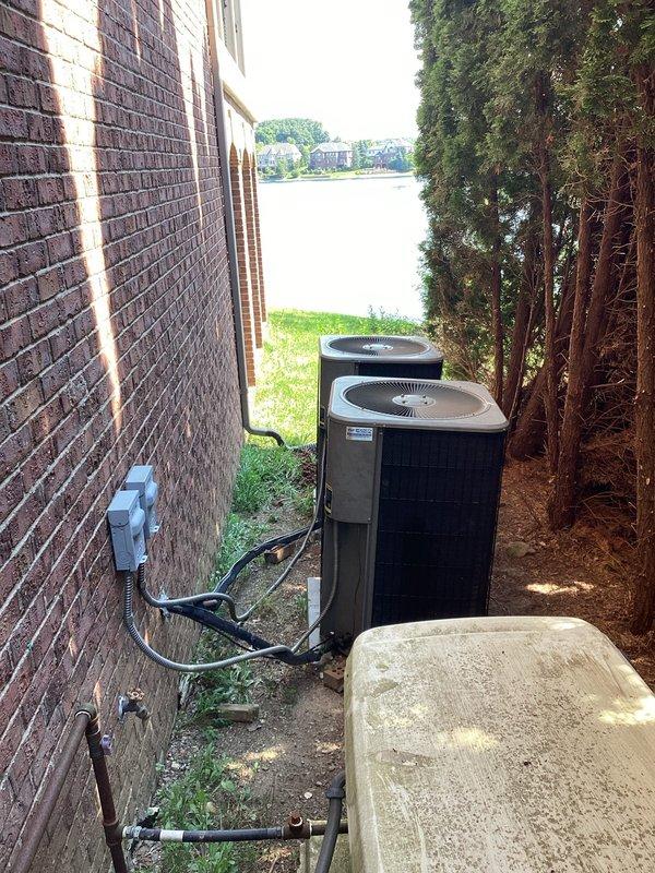 Completed on-site evaluation for air conditioning system replacement. Assessed existing dual outdoor condenser units located in side yard between brick structure and evergreen hedge, documented current equipment condition showing weathering and dirt accumulation on mesh grilles, photographed electrical conduit routing along brick wall and refrigerant line configurations, and evaluated installation space constraints in narrow access area with concrete pad foundation. Prepared comprehensive replacement estimate based on site conditions and equipment specifications.