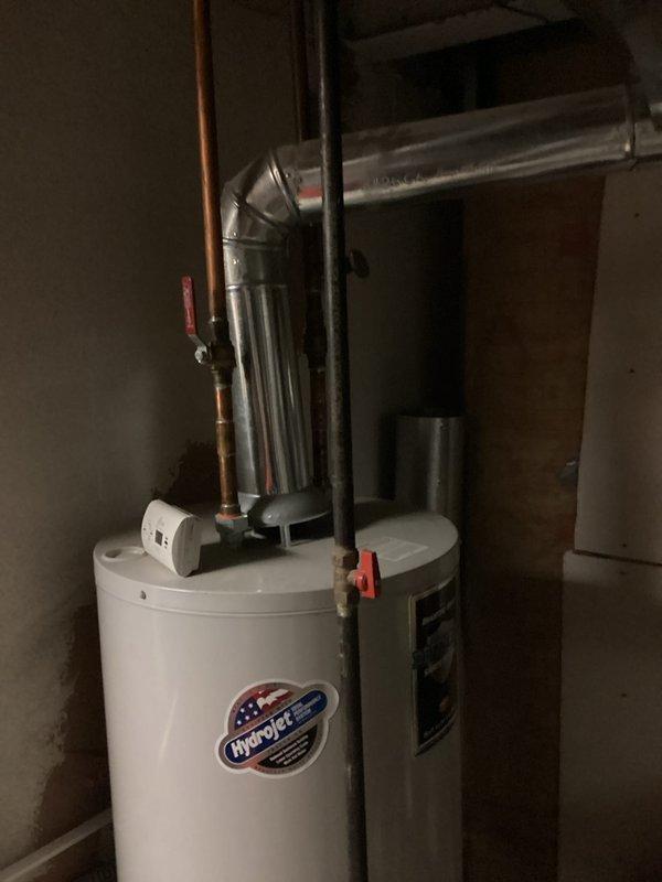 Responded to service call for water heater leaking from bottom in basement utility area. Upon arrival, conducted thorough inspection of white cylindrical residential water heater with visible copper supply lines and discharge piping. Documented existing conditions through photography showing unit positioned adjacent to HVAC equipment with metallic ductwork overhead. Visual examination revealed active leak originating from lower section of tank, indicating potential internal tank failure or deterioration of bottom drain valve assembly. Assessed surrounding area for water damage and confirmed concrete floor showed signs of moisture accumulation. Evaluated age and condition of unit based on manufacturer label and overall system configuration. Photographed installation setup including copper piping connections, pressure relief valve, and gas supply lines for estimate documentation. Provided comprehensive estimate for water heater replacement including removal of existing failed unit, prope