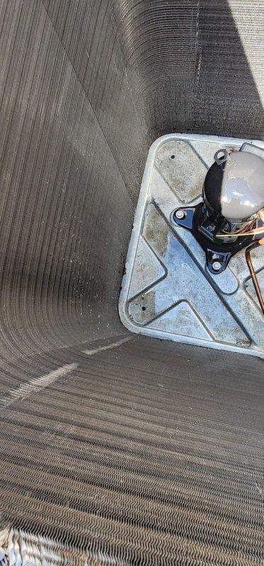 Completed routine maintenance on outdoor air conditioning unit and heat pump system. Inspected cylindrical condenser unit and documented significant accumulation of organic debris, dirt, and leaves along the top edge and between the horizontal metal fins. Debris buildup was particularly heavy in the upper rim area, indicating need for thorough cleaning to maintain optimal heat exchange efficiency. Examined interior air handler compartment and found extensive dirt and particulate matter coating interior surfaces with visible striations throughout. Verified compressor unit securely mounted with proper bracket installation and confirmed refrigerant lines properly connected. Unit operational but requires cleaning to restore full efficiency and prevent potential airflow restrictions that could impact system performance.