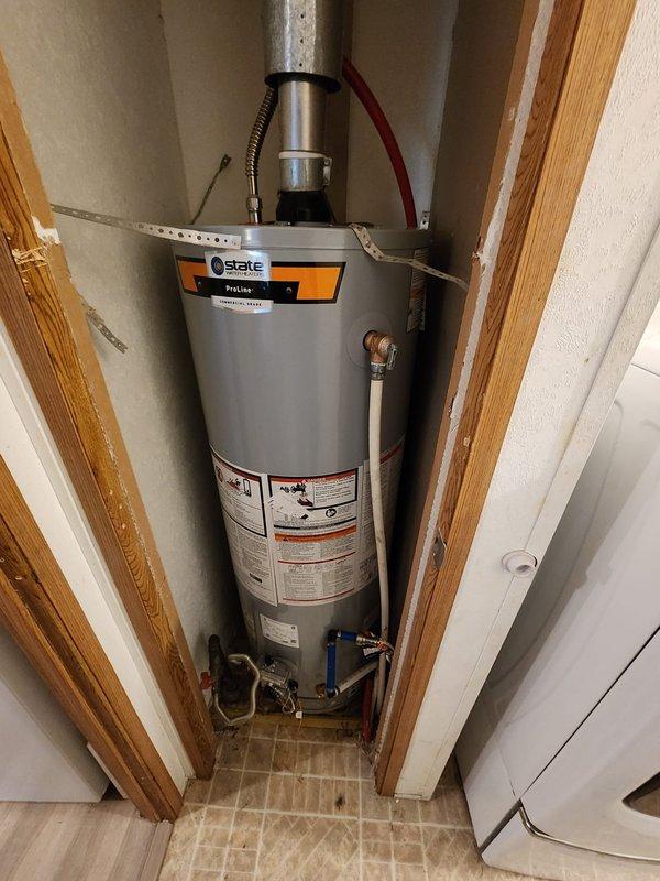 Completed installation of a 40-gallon State ProLine tanked water heater in a mobile home utility closet. Positioned unit on yellow plastic drain pan over herringbone-patterned tile flooring and secured to wall with metal strapping for seismic safety. Connected water supply lines with blue shut-off valves, installed pressure relief valve with discharge piping, and completed electrical connections using flexible conduit. Verified all plumbing connections for leaks, tested unit operation, and confirmed proper venting through existing exhaust system.