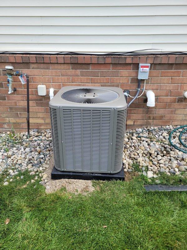 Successfully installed a new Lennox central air conditioning condenser unit in the residential backyard, positioned on a concrete pad with proper base platform and river rock landscaping for drainage. The installation included complete refrigerant line connections with protective sleeves, electrical disconnect box mounting to the brick exterior wall with PVC conduit runs, and all necessary piping connections to integrate with the existing HVAC system.