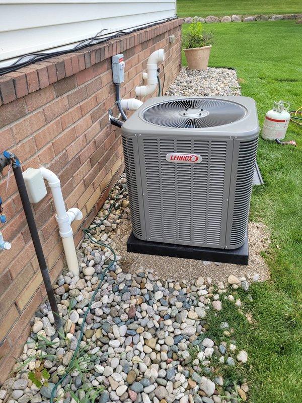 Successfully installed a new Lennox central air conditioning condenser unit in the residential backyard, positioned on a concrete pad with proper base platform and river rock landscaping for drainage. The installation included complete refrigerant line connections with protective sleeves, electrical disconnect box mounting to the brick exterior wall with PVC conduit runs, and all necessary piping connections to integrate with the existing HVAC system.