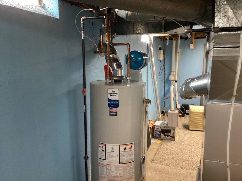 Completed installation of a 50-gallon Bradford White tanked water heater in basement utility room. Connected copper supply and discharge piping to unit with proper shutoff valves and fittings, installed blue expansion tank on wall to manage thermal expansion in closed plumbing system, and ran black TPR discharge pipe vertically along side of unit per code requirements. All connections pressure tested and water heater commissioned successfully with proper venting configuration confirmed.