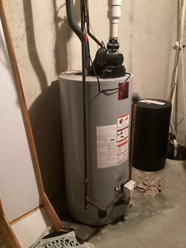 Conducted comprehensive on-site evaluation for hot water heater replacement in residential basement utility area. Documented existing gray cylindrical water heater installation, including current plumbing connections with black and copper piping, white PVC venting system, pressure tank configuration, and electrical connections. Assessed overhead structural elements, exposed joists, and existing drain/vent pipe routing to determine optimal replacement specifications and installation requirements.