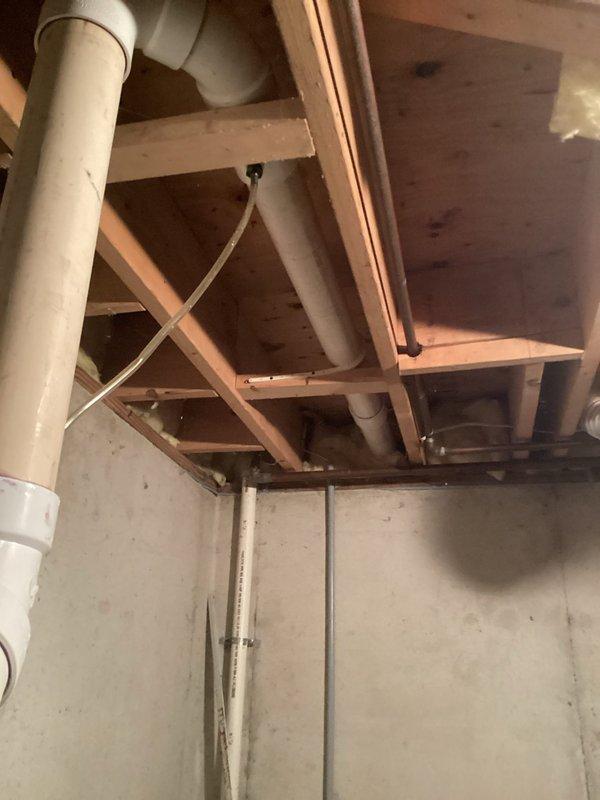 Conducted comprehensive on-site evaluation for hot water heater replacement in residential basement utility area. Documented existing gray cylindrical water heater installation, including current plumbing connections with black and copper piping, white PVC venting system, pressure tank configuration, and electrical connections. Assessed overhead structural elements, exposed joists, and existing drain/vent pipe routing to determine optimal replacement specifications and installation requirements.