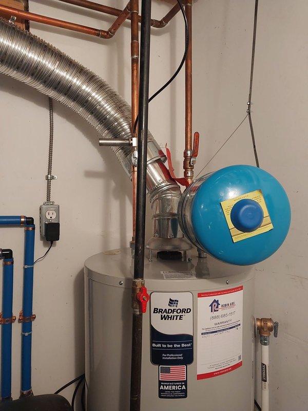 Completed warranty inspection and corrections on Bradford White water heater installation. Addressed three code compliance issues: repositioned temperature and pressure relief valve to provide adequate clearance from wall for proper operation and future replacement access, installed required safety drain pan beneath water heater per Section 504.7 through 504.7.2 of the 2021 Michigan Plumbing Code, and secured flexible vent connections to water heater and chimney with appropriate screws. Documentation photos captured of corrected installation showing properly positioned relief valve, compliant drain pan installation, and secured vent connections along with associated copper piping, blue expansion tank, and mechanical room components.