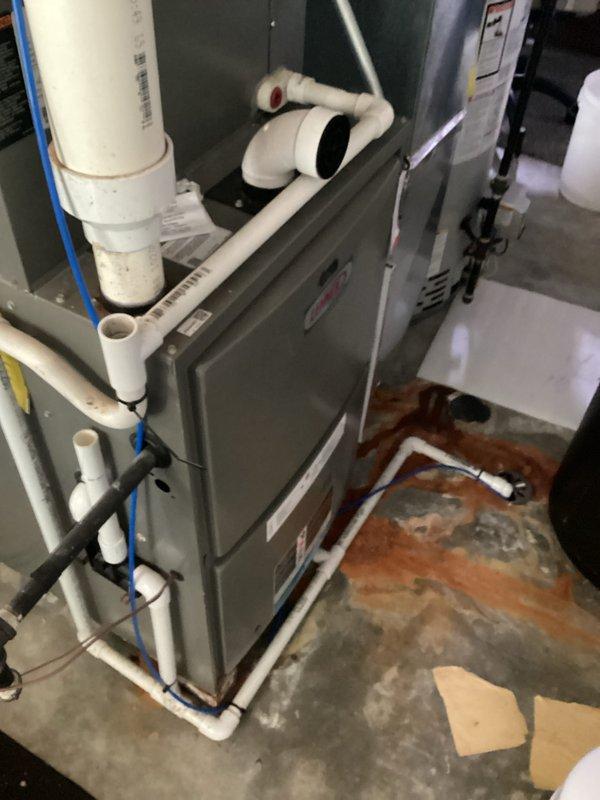 Conducted comprehensive furnace replacement estimate for residential HVAC system with failed heat exchanger. Documented existing gray metal air handler installation showing significant water damage on surrounding concrete floor with rust-colored staining, indicating previous moisture intrusion or condensate drainage issues. Photographed complete system configuration including white PVC condensate piping, blue wiring, electrical control panel with yellow manufacturer label, green cylindrical air handler unit, and associated plumbing connections with red valve handle on gray supply lines. Assessed mechanical room conditions and equipment accessibility for replacement procedures.