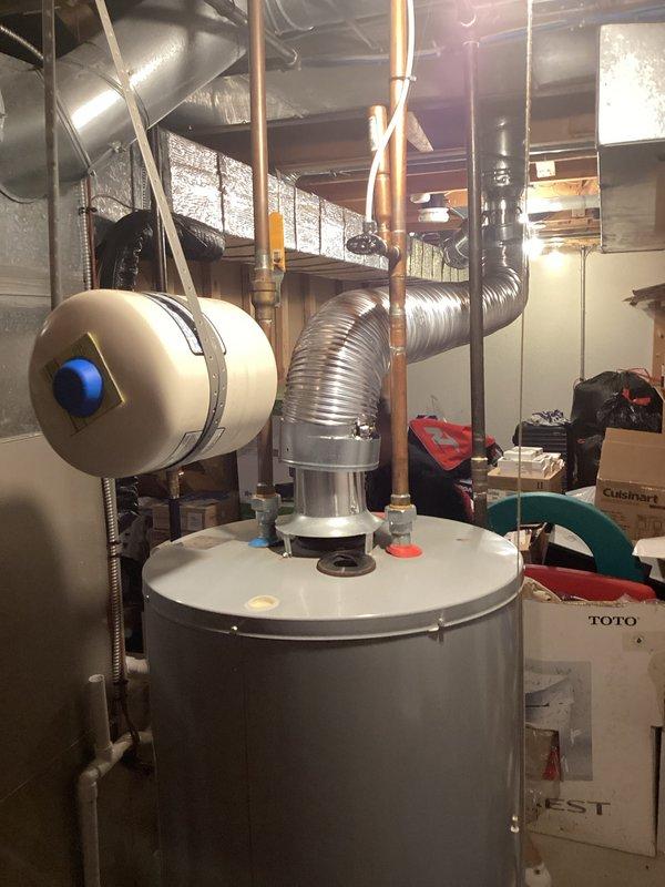 Completed estimate for water heater replacement in residential basement utility room. Existing gray cylindrical water heater showing signs of base leakage with visible water staining on concrete floor surrounding unit. Assessed current installation including gas connections, plumbing infrastructure, venting system with aluminum flexible ductwork, and proximity to existing HVAC equipment including humidifier unit. Documented equipment specifications, clearances, and existing piping configuration to provide accurate replacement proposal.