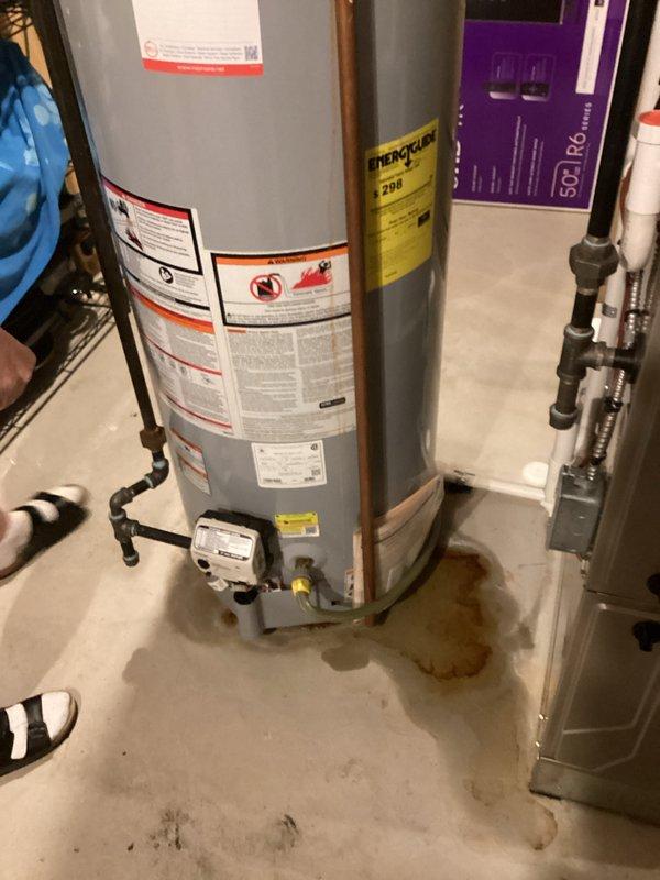 Completed estimate for water heater replacement in residential basement utility room. Existing gray cylindrical water heater showing signs of base leakage with visible water staining on concrete floor surrounding unit. Assessed current installation including gas connections, plumbing infrastructure, venting system with aluminum flexible ductwork, and proximity to existing HVAC equipment including humidifier unit. Documented equipment specifications, clearances, and existing piping configuration to provide accurate replacement proposal.