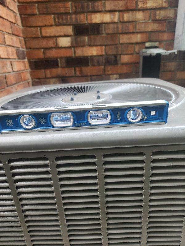 Responded to callback for outdoor AC condenser unit evaluation. Upon arrival, assessed unit for noise concerns during startup cycle and verified levelness of installation. Used precision level to check condenser positioning on mounting pad and concrete base. Unit was found to be properly leveled with no adjustment required. Conducted startup observation and monitored unit operation through multiple cycles between 8-12 timeframe as specified. No abnormal sounds or vibrations detected during compressor engagement or fan motor operation. Condenser operating within normal parameters with proper airflow through louvered panels and fan assembly functioning correctly. Installation remains secure on rubber mounting pad with adequate clearance from brick structure. Documented current condition with photos showing proper level placement and overall unit positioning for service records.