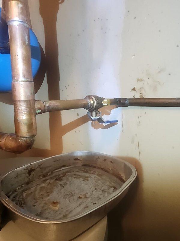 Responded to water leak at tanked water heater installation. Upon inspection, identified active leak at soldered connection on 1/2-inch ball valve with bleeder drain. The copper piping system showed signs of age and weathering, with visible corrosion and patina on connections. Repaired faulty solder joint at valve connection point, ensuring proper seal and eliminating leak. Inspected drain pan beneath water heater, which showed evidence of previous water accumulation and rust staining. Tested all connections for integrity after repair and verified system was operating without leaks.