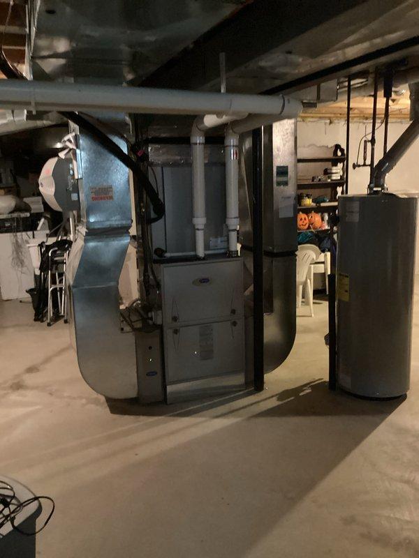 Conducted comprehensive assessment of existing HVAC system in residential basement mechanical room for equipment replacement estimate. Documented current furnace unit with connected ductwork, water heater installation, and associated mechanical systems including white PVC condensate drainage lines and ventilation piping. Evaluated equipment placement, existing duct configuration with both rigid and flexible metallic ductwork connections, and available workspace for potential system replacement. Photographed basement utility area showing concrete floor installation, exposed ceiling joists, and current equipment positioning to provide accurate replacement specifications and installation planning requirements.