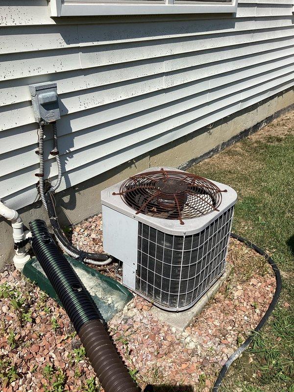 Performed site evaluation for 27-year-old residential air conditioning system replacement. The existing outdoor condensing unit showed signs of age-related failure and was no longer operational. Documented current equipment configuration including outdoor condenser unit positioned on concrete pad with decorative rock landscaping, refrigerant line set routing through black flexible conduit, and indoor air handler/furnace system located in basement utility area. Photographed existing installation showing proper clearances, electrical connections, and ductwork connections to facilitate accurate equipment sizing and replacement planning for new HVAC system installation.