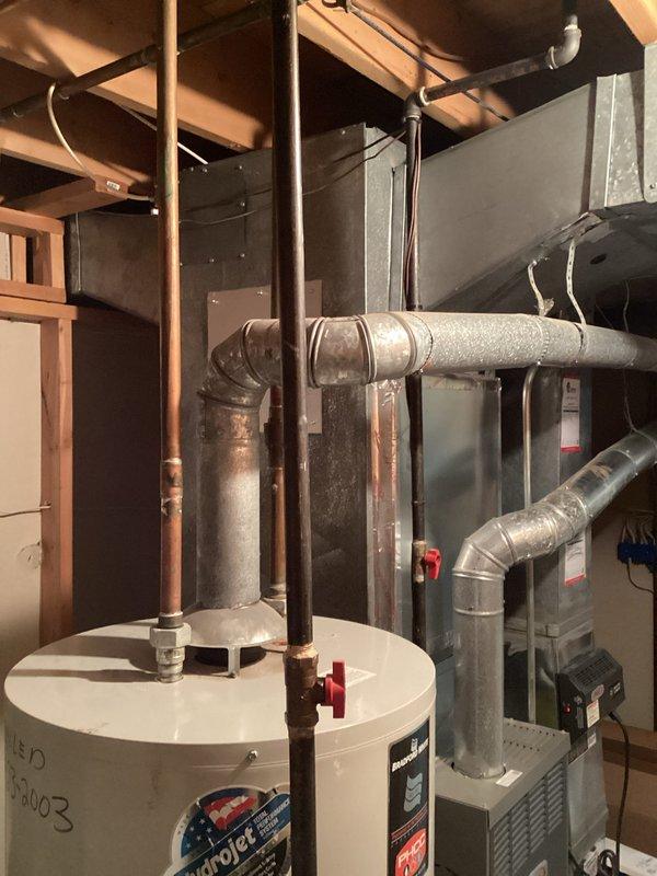 Conducted on-site assessment and provided estimate for water heater replacement in residential basement mechanical room. Evaluated existing white cylindrical tank-style water heater with visible manufacturer specifications and aging components, including copper supply piping with red valve handles. Documented current installation configuration adjacent to gray furnace unit with metallic ductwork in unfinished utility space. Photographed equipment conditions, piping connections, venting arrangements, and clearances to nearby HVAC system for accurate replacement planning and material specifications.